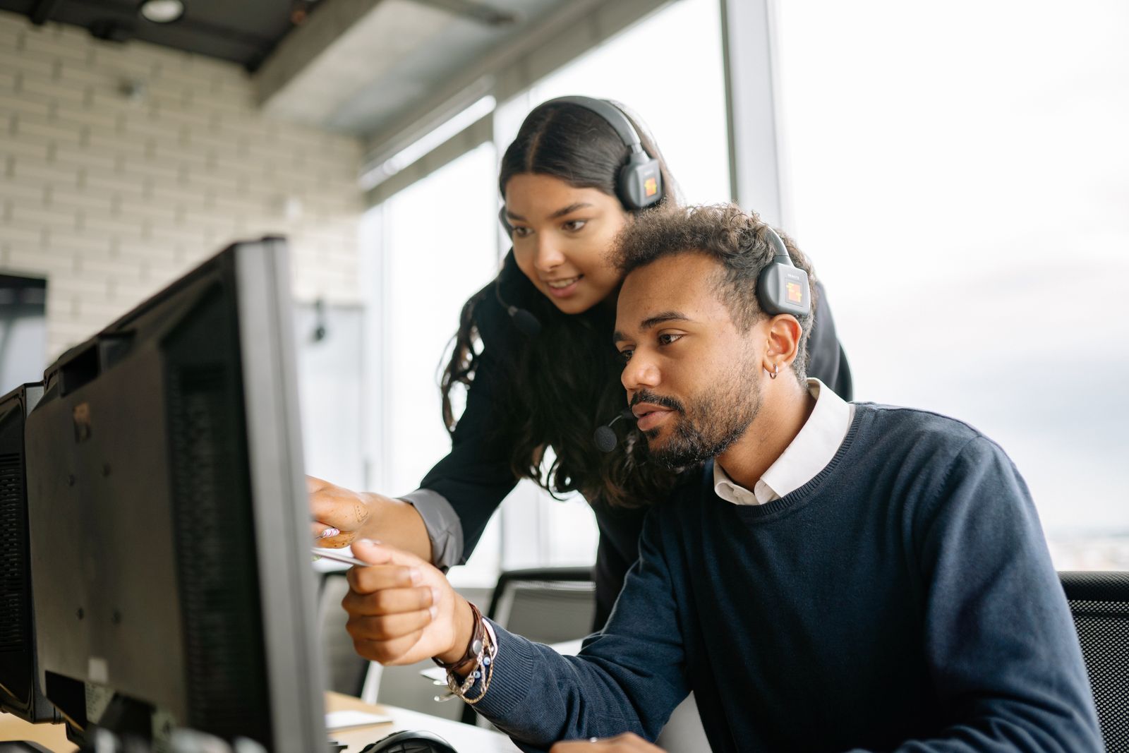 Mentor coaching a new contact center agent in real time during nesting, leaning over their shoulder and pointing at the screen mid-call